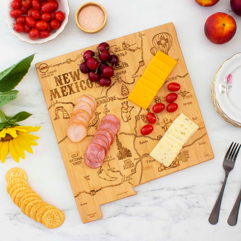 The Destination New Mexico Cutting Board by Totally Bamboo, measuring 11-3/4" x 13-1/2" and crafted from bamboo, is adorned with sliced meats, cheeses, tomatoes, and grapes. Nearby on the light marble surface are a fork, crackers, sunflowers, and peaches. A decorative plate is also present.