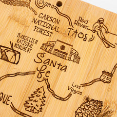 Introducing the Destination New Mexico Cutting Board by Totally Bamboo, measuring 11-3/4" x 13-1/2". This bamboo board is laser-engraved with a detailed map highlighting Santa Fe, Taos, Carson National Forest, Bandelier National Monument, Red River, Chama, Las Vegas, and a skier. Charming illustrations of a tree and pine cone add to its appeal.