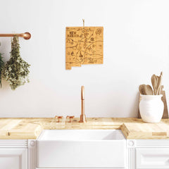 A kitchen showcasing a white sink and wooden countertop is highlighted by a copper faucet centrally placed on the counter, accompanied by hanging greenery to the left. Above the sink, a Totally Bamboo Destination New Mexico Cutting Board, featuring laser-engraved artwork of New Mexico, adds a distinctive touch to the decor.