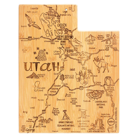 The "Destination Utah Cutting Board" by Totally Bamboo is an 11" x 14-1/4" laser-engraved board shaped like the state of Utah, featuring intricate illustrations of cities and landmarks like Salt Lake City, Provo, and Arches National Park, as well as depictions of wildlife and mountains on a bamboo cutting surface.