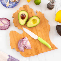 The Destination Michigan Mitten Cutting Board by Totally Bamboo, measuring 13-1/4" x 11-3/4", elegantly holds a sliced avocado and red onion. Nearby, a green-handled knife is accompanied by a lemon, lime, and black pepper grinder. This bamboo board is complemented by a decorative plate with a fork, adding charm to your kitchen setting.