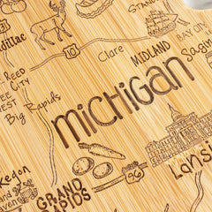 The Totally Bamboo Destination Michigan Mitten Cutting Board, measuring 13-1/4" x 11-3/4", is a state-shaped wooden map resembling Michigan. It features laser-engraved artwork that highlights cities such as Clare, Lansing, and Grand Rapids with illustrations of ships and buildings. The board also includes highway numbers 10 and 96 to emphasize significant locations and landmarks.