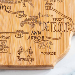 The Totally Bamboo Destination Michigan Mitten Cutting Board, measuring 13-1/4" x 11-3/4", showcases a map with select Michigan locations like Detroit, Ann Arbor, and Monroe. It features laser-engraved artwork adorned with vintage cars and flags.