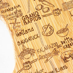 The Totally Bamboo Destination Michigan Mitten Cutting Board, measuring 13-1/4" x 11-3/4", features a laser-engraved depiction of Michigan cities such as Grand Rapids, Muskegon, and Kalamazoo, embellished with illustrations of local icons like an airplane, paint palette, and a bowl of food. This intricate design is crafted on a textured wooden surface.