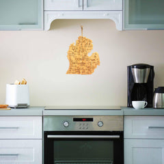 In a modern kitchen featuring white cabinets and silver appliances, a coffee maker, toaster, and cup rest on the countertop. A Destination Michigan Mitten Cutting Board from Totally Bamboo hangs on the beige wall above the stove, measuring 13-1/4" x 11-3/4".