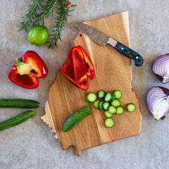 Arranged on the Rock & Branch® Origins Series Georgia Cutting Board by Totally Bamboo, you’ll find sliced cucumbers, a whole cucumber, half a red bell pepper, purple onion, rosemary, and a knife. Lying on the gray surface nearby are a lime, another red bell pepper, and an additional cucumber.