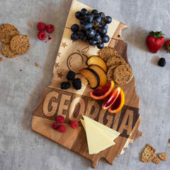 The Rock & Branch® Origins Series Georgia Cutting Board by Totally Bamboo, crafted from rich acacia wood and laser-engraved in the shape of Georgia, beautifully cradles a delightful array: grapes, plums, oranges, brie slices, raspberries, blackberries, strawberries, and whole-grain crackers.