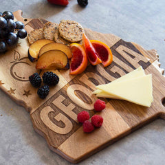 The Rock & Branch® Origins Series Georgia Cutting Board by Totally Bamboo is a state-shaped acacia wood board, beautifully laser-engraved with "Georgia" and a peach design. It elegantly displays sliced cheese, crackers, blackberries, raspberries, grapes, peach slices, and orange slices.