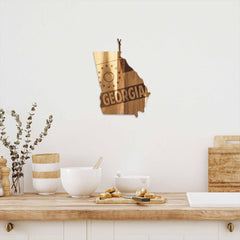 A cozy kitchen scene is enhanced by the Rock & Branch® Origins Series Georgia Cutting Board from Totally Bamboo, elegantly crafted from acacia wood and laser-engraved in the shape of Georgia with the state's name. Below, a countertop showcases bowls, other cutting boards, and pastries, while a small vase with greenery provides a natural touch.