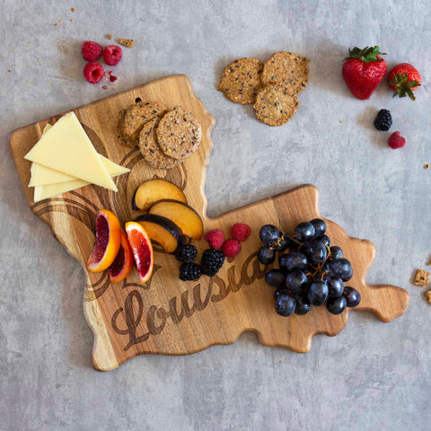 The Rock & Branch® Origins Series Louisiana Cutting Board by Totally Bamboo, crafted from acacia wood, elegantly displays slices of cheese, crackers, peach slices, citrus halves, blackberries, raspberries, strawberries, and a bunch of grapes on a gray surface. The laser-engraved artwork captures the essence of Louisiana.