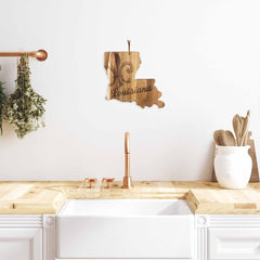 The kitchen has a warm atmosphere, highlighted by the Rock & Branch® Origins Series Louisiana Cutting Board from Totally Bamboo, laser-engraved with a fleur-de-lis and the word "Louisiana." The room is enhanced by an acacia wood countertop and features a white sink paired with a copper faucet. Nearby, herbs are neatly hung for easy access. To the right, utensils are organized in a clay pot.