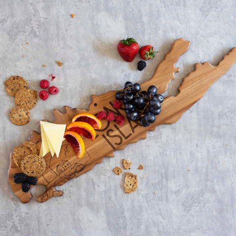 The Rock & Branch® Origins Series Long Island Cutting Board from Totally Bamboo, crafted from acacia wood and shaped like an island, features grapes, cheese slices, orange segments, berries, and crackers. On the surrounding gray surface lie scattered crackers and berries. Near the board's top edge rest two strawberries.