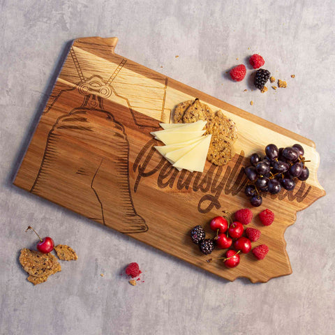 The Rock & Branch® Origins Series Pennsylvania Cutting Board by Totally Bamboo features laser-engraved Liberty Bell artwork. Crackers, cheese slices, grapes, cherries, and raspberries are beautifully arranged on this acacia wood masterpiece set against a gray backdrop with scattered crackers.