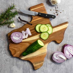 The Rock & Branch® Origins Series Texas Cutting Board by Totally Bamboo, crafted from acacia wood and shaped like the state of Texas, displays sliced cucumbers, a partially sliced cucumber, pieces of red onion, and a knife. Whole garlic cloves and rosemary rest nearby on the gray surface, adding a touch of rustic charm to this unique cutting board.