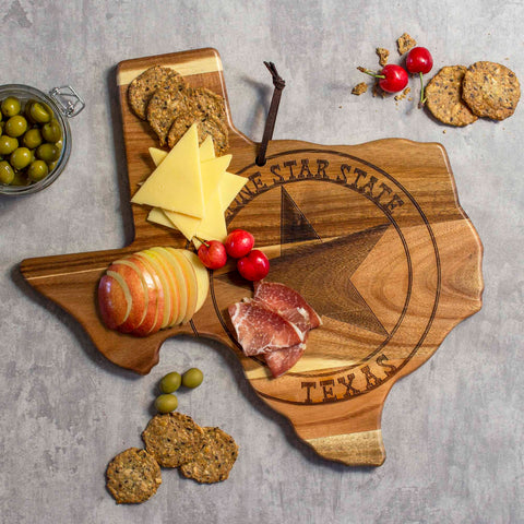 The Rock & Branch® Origins Series Texas Cutting Board by Totally Bamboo, crafted from acacia wood, elegantly displays an array of sliced cheese, apples, cherry tomatoes, and cured meats. Crackers are artfully arranged within the spread. Nearby on a textured surface sits a small jar of olives, enhancing the rustic charm.