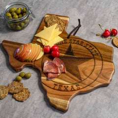 The Rock & Branch® Origins Series Texas Cutting Board by Totally Bamboo, crafted from acacia wood, features a laser-engraved star design and "Lone Star State Texas" artwork. It is beautifully adorned with cheese slices, apple slices, cured meat, and cherries, while olives fill a nearby jar to complete the presentation.