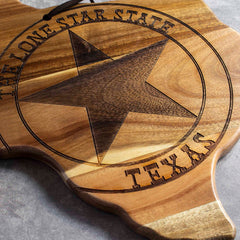 The Rock & Branch® Origins Series Texas Cutting Board by Totally Bamboo, made from acacia wood in the iconic shape of Texas, showcases a laser-engraved star encircled by "The Lone Star State Texas" at its center. The board rests gracefully on a gray surface.