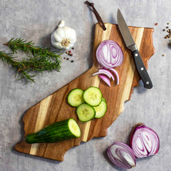 On a Rock & Branch® Origins Series California Cutting Board by Totally Bamboo, sliced cucumbers and onions are arranged like a charcuterie board, with a knife resting on the wooden surface. On the concrete surface nearby, there is a whole cucumber, halved onion, garlic bulb, and rosemary sprigs.