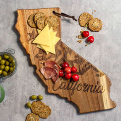 A Rock & Branch® Origins Series California Cutting Board by Totally Bamboo, crafted from acacia wood and adorned with laser-engraved "California" and bear artwork, stylishly holds cheese, crackers, meats, and cherries. It rests on a gray surface next to a jar of green olives.