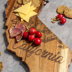 The Rock & Branch® Origins Series California Cutting Board by Totally Bamboo, crafted from acacia wood and featuring laser-engraved artwork, holds sliced cheese, prosciutto, cherries, and scattered crackers on a gray surface.