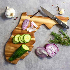 A Rock & Branch® Origins Series Florida Cutting Board by Totally Bamboo, crafted from Acacia wood and shaped like the state of Florida, holds sliced cucumbers and red onions. A knife lies on the board. On the gray stone surface are garlic, cracked black pepper, rosemary, and a halved red onion—each detail highlighted by laser-engraved artwork.