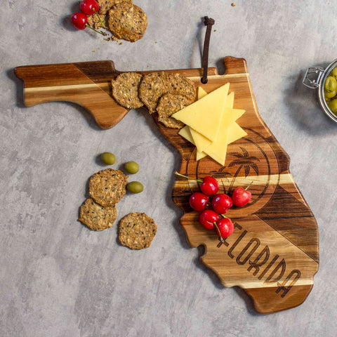 The Rock & Branch® Origins Series Florida Cutting Board by Totally Bamboo, crafted from Acacia wood, showcases crackers, cheese slices, and cherry tomatoes. A laser-engraved "Florida" adorns the state-shaped board. Adding to this charming presentation are additional crackers and green olives nearby.