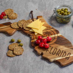 A Rock & Branch® Origins Series Florida Cutting Board by Totally Bamboo, crafted from acacia wood and shaped like the state of Florida, elegantly displays sliced cheese, cherry tomatoes, and crackers. Nearby sits a small jar of green olives. The board features the word "Florida" laser-engraved with finesse on its surface.