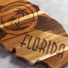 The Rock & Branch® Origins Series Florida Cutting Board by Totally Bamboo is crafted from acacia wood in the shape of Florida, featuring laser-engraved palm trees within a circle and "FLORIDA" in bold letters. The natural wood grain displays an array of shades that enhance its distinct appeal.