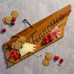 The Rock & Branch® Origins Series Tennessee Cutting Board by Totally Bamboo, made from acacia wood, is designed in the shape of Tennessee and beautifully holds crackers, cheese slices, prosciutto, and cherries. The gray surface around this delightful setup features scattered fresh berries and additional crackers.