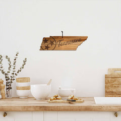 A kitchen countertop features bowls, a small potted plant, and a wooden cutting board crafted from Acacia wood. Hanging on the wall above is the Rock & Branch® Origins Series Tennessee Cutting Board by Totally Bamboo, shaped like Tennessee with "Tennessee" and stars laser-engraved on it.