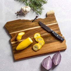 The Rock & Branch® Origins Series Oregon Cutting Board by Totally Bamboo, crafted from acacia wood, holds sliced yellow squash with a knife resting beside it. Nearby, a halved red onion, garlic bulb, peppercorns, and rosemary sprigs are artistically arranged on a light gray surface.