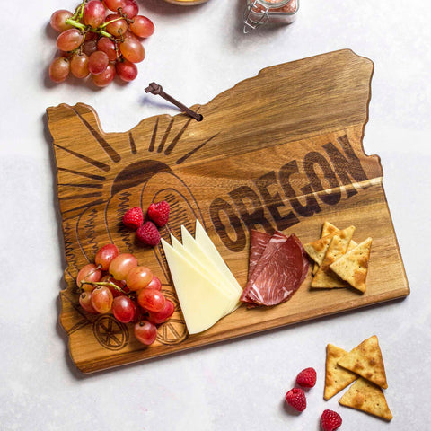 The Rock & Branch® Origins Series Oregon Cutting Board by Totally Bamboo, made from acacia wood and laser-engraved with "Oregon" and a covered wagon design, elegantly displays grapes, raspberries, cheese slices, cured meats, and crackers on a light background decorated with scattered raspberries and crackers.