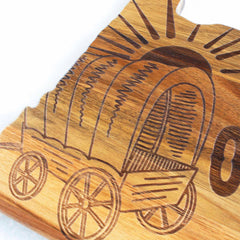 The Rock & Branch® Origins Series Oregon Cutting Board by Totally Bamboo is designed to emulate a covered wagon with rays of light reaching upward. This acacia wood cutting board showcases four wheels and detailed carvings, evoking a sense of motion and an antique style.