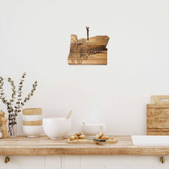 On a kitchen countertop with white bowls and wooden utensils, there's a small Rock & Branch® Origins Series Oregon Cutting Board by Totally Bamboo hanging on the white wall, featuring a laser-engraved wagon design. Decorative branches in a vase are placed on the left.