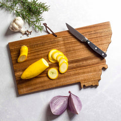 The Rock & Branch® Origins Series Montana Cutting Board by Totally Bamboo showcases a paring knife, sliced yellow squash, garlic, rosemary, and peppercorns. Nearby, half of a red onion rests on the white marble surface, crafting a Montana-themed artwork for your kitchen.