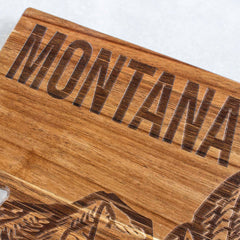 The Rock & Branch® Origins Series Montana Cutting Board by Totally Bamboo showcases "MONTANA" beautifully engraved at the top, complemented by an intricate mountain carving beneath. Made of acacia wood, it features a rich natural color and distinctive grain patterns, capturing state-themed artistry in each polished curve.