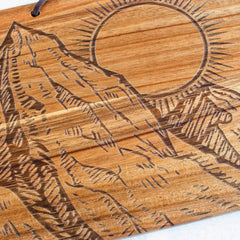 The Rock & Branch® Origins Series Montana Cutting Board by Totally Bamboo features an engraved depiction of a stylized mountain range on acacia wood, accented with a large sun encircled by rays in the upper right corner. This state-themed cutting board exudes a natural, rustic charm that beautifully captures the essence of regional beauty.