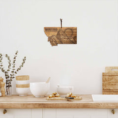 A kitchen interior showcases a Montana-shaped wall decoration crafted from acacia wood, adorned with mountain and sun engravings. Positioned on the countertop is the Rock & Branch® Origins Series Montana Cutting Board by Totally Bamboo, alongside white bowls, a plant, and a few croissants on a tray, creating a cozy, rustic atmosphere.