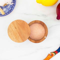 The Rock & Branch® Salt Cellar from Totally Bamboo, featuring a 6-ounce capacity and a secure magnetic lock, is filled with pink Himalayan salt on a marble surface. Surrounding it are a garlic bulb, yellow lemon, red onion, colorful spoon, and decorative blue plate that enhance the vibrant display.