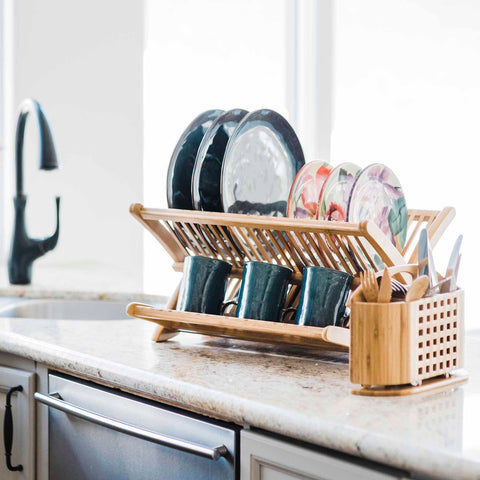 A wooden Eco Dish Rack on a kitchen countertop holds a mix of dark and floral-patterned plates along with several blue mugs. To the right, the Utensil Caddy for Eco Dish Rack by Totally Bamboo elegantly cradles forks and knives. A sink with a modern faucet gleams in the background, completing the harmonious kitchen scene.