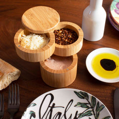 The Totally Bamboo Triple Salt Cellar with Swivel Lids, boasting a 1-1/3 cups capacity, elegantly displays grated cheese, crushed red pepper, and salt in its three open compartments. It enhances the dining table setting alongside fresh bread, a decorative plate, and olive oil complemented by balsamic vinegar. Its swivel lids ensure effortless access.