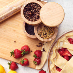 Top-down view of a kitchen scene featuring fresh strawberries, lemons, a rolling pin, and the Totally Bamboo Triple Salt Cellar with Swivel Lids filled with star anise, allspice, poppy seeds, and cardamom. A partially made pie with a crisscross top and a wooden cutting board are also visible.