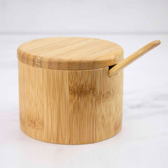 The Totally Bamboo Little Dipper Salt Cellar, featuring a 6-ounce capacity, rests on a white surface. A matching lid accompanies it, and a small wooden spoon is nestled in a notch, suggesting the lid can close with the spoon inside. The background is softly blurred.