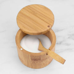 A Little Dipper Salt Cellar with a swiveling lid from Totally Bamboo, filled with sugar, sits on a marble surface. The lid is partially open, and the included salt spoon rests inside, protruding over the edge.