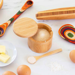 A Totally Bamboo Little Dipper Salt Cellar with a salt spoon, featuring a 6-ounce capacity, sits on a marble surface. It is surrounded by colorful measuring spoons, a wooden rolling pin, a plate of butter, and eggs.