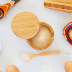 A Little Dipper Salt Cellar with Salt Spoon from Totally Bamboo is positioned on a white surface, filled with sugar and accompanied by vibrant wooden spoons, a wooden butter dish, eggs, and scattered flour. A small wooden spoon holds a portion of sugar, while the magnetic lock ensures the sweetness stays secure.