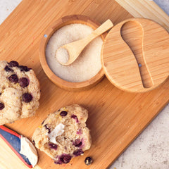 A wooden board displays two blueberry muffins, one cut in half, next to the Totally Bamboo Big Dipper Salt Cellar with a salt spoon. A knife with a blue handle and some butter rest nearby, highlighting a sense of rustic elegance.