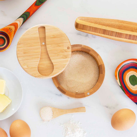 On a white countertop sits the Big Dipper Salt Cellar with a salt spoon by Totally Bamboo, filled with sugar. Nearby are wooden kitchen utensils and radiant handles surrounding a plate of butter and raw eggs. A small wooden spoon with sugar completes the scene.