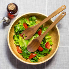 A 12" TB Home® Acacia Wood Salad Serving Set by Totally Bamboo showcases a wooden bowl brimming with a fresh green salad of chopped lettuce and tomato wedges. The set includes acacia wood salad servers resting on top, while nearby, an oil bottle and pepper shaker sit on a gray tablecloth, perfectly completing this elegant arrangement.