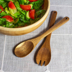 A wooden salad bowl filled with lettuce, sliced tomatoes, and cucumbers is complemented by the TB Home® Acacia Wood Salad Serving Set by Totally Bamboo. The elegant 12" utensils are artfully arranged beside the bowl on a gray cloth surface.